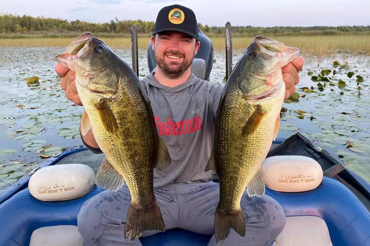 An angler sitting in a boat holding up two largemouth bass; the boat is in a field of lily pads.