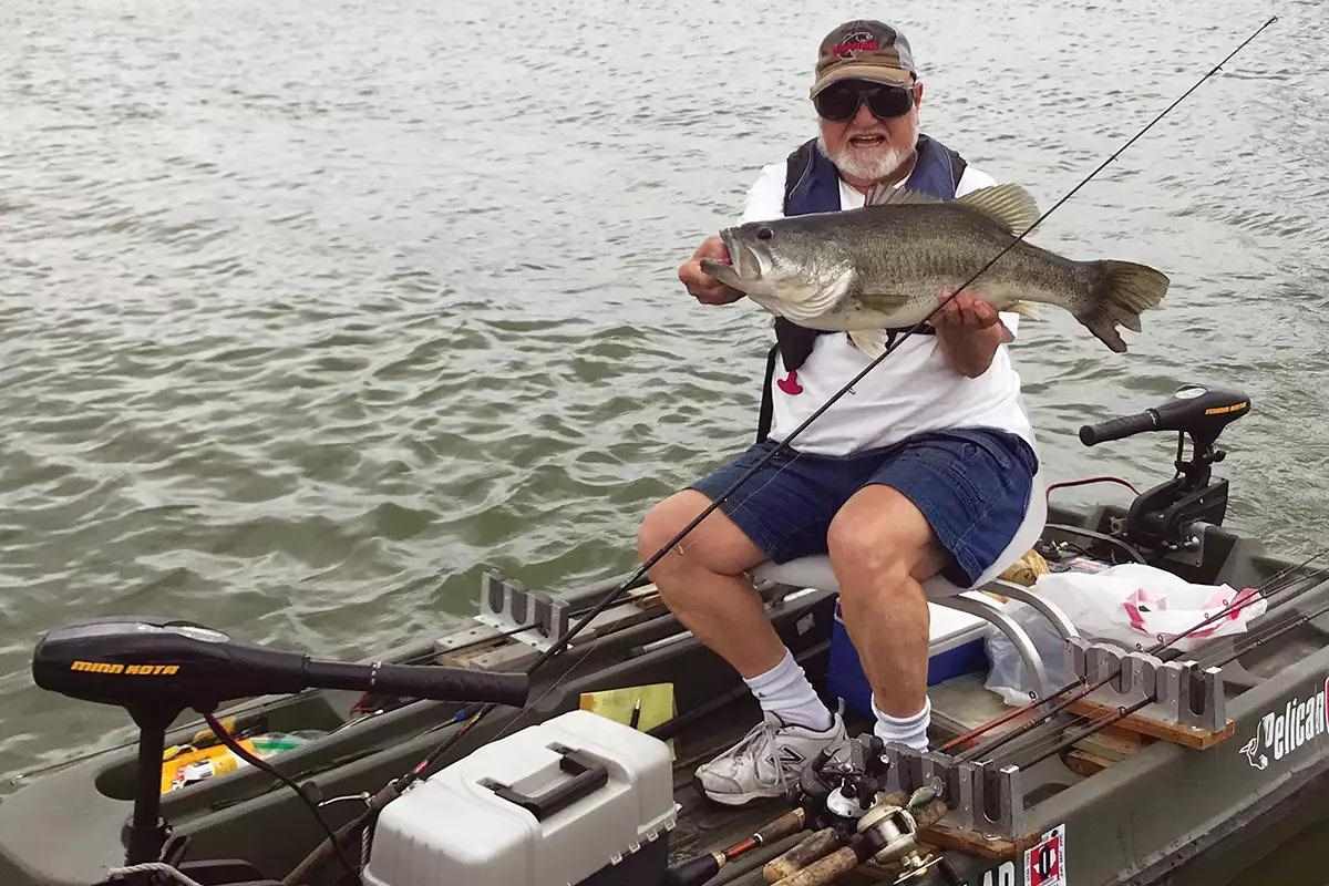 A bearded fisherman sitting in a small boat holding a large guadalupe bass. 