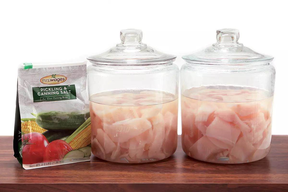 Two lidded jars with fish fillets and vinegar next to a bag of pickling salt on a countertop.